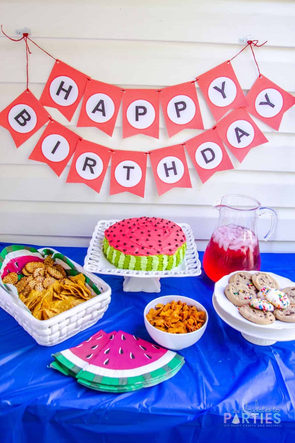 angled down photo of a party table and banner with birthday cake and snacks