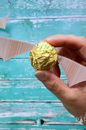 A woman's hand holding a Ferrero Rocher truffle with wings added to look like a Harry Potter golden snitch.