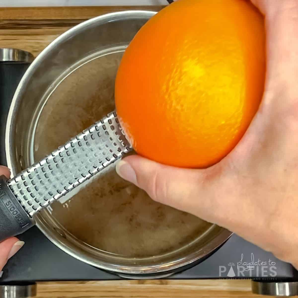 A woman's hand using a microplane to zest an orange into a pot of liquid.