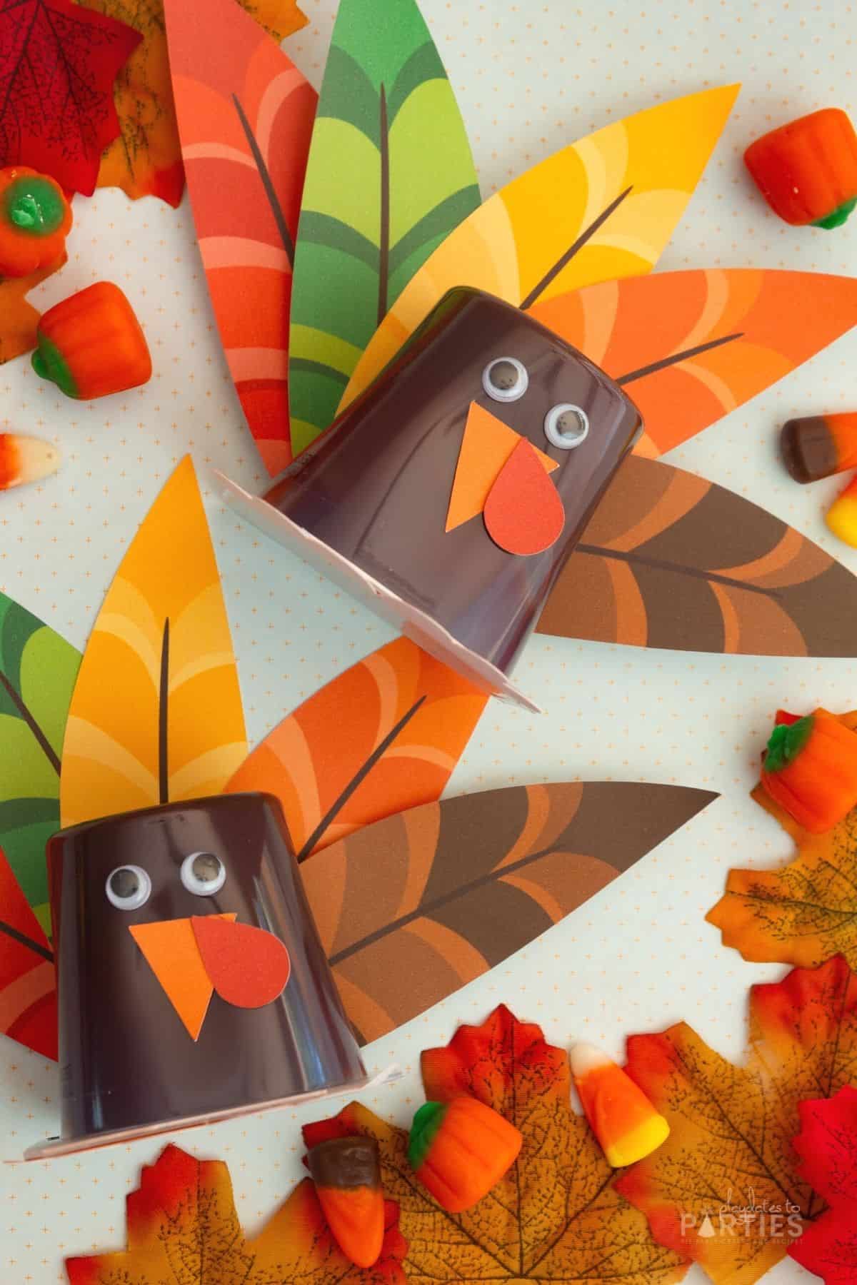 Two turkey pudding cups laying on a piece of blue paper with orange dots, surrounded by leaves and candy corn.