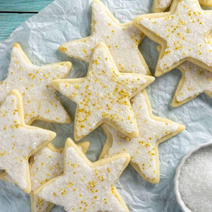 Sugar cookies decorated like stars on a blue wood surface with a bowl of sugar crystals nearby.