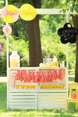 A white wood lemonade stand with pink decorations sitting in a green park.