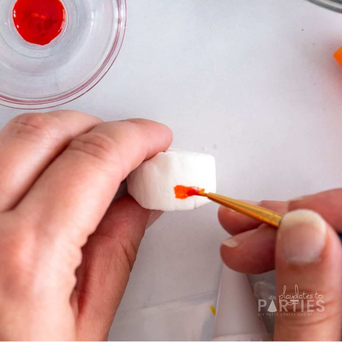 A woman's hand painting an orange carrot nose onto a marshmallow with food coloring.