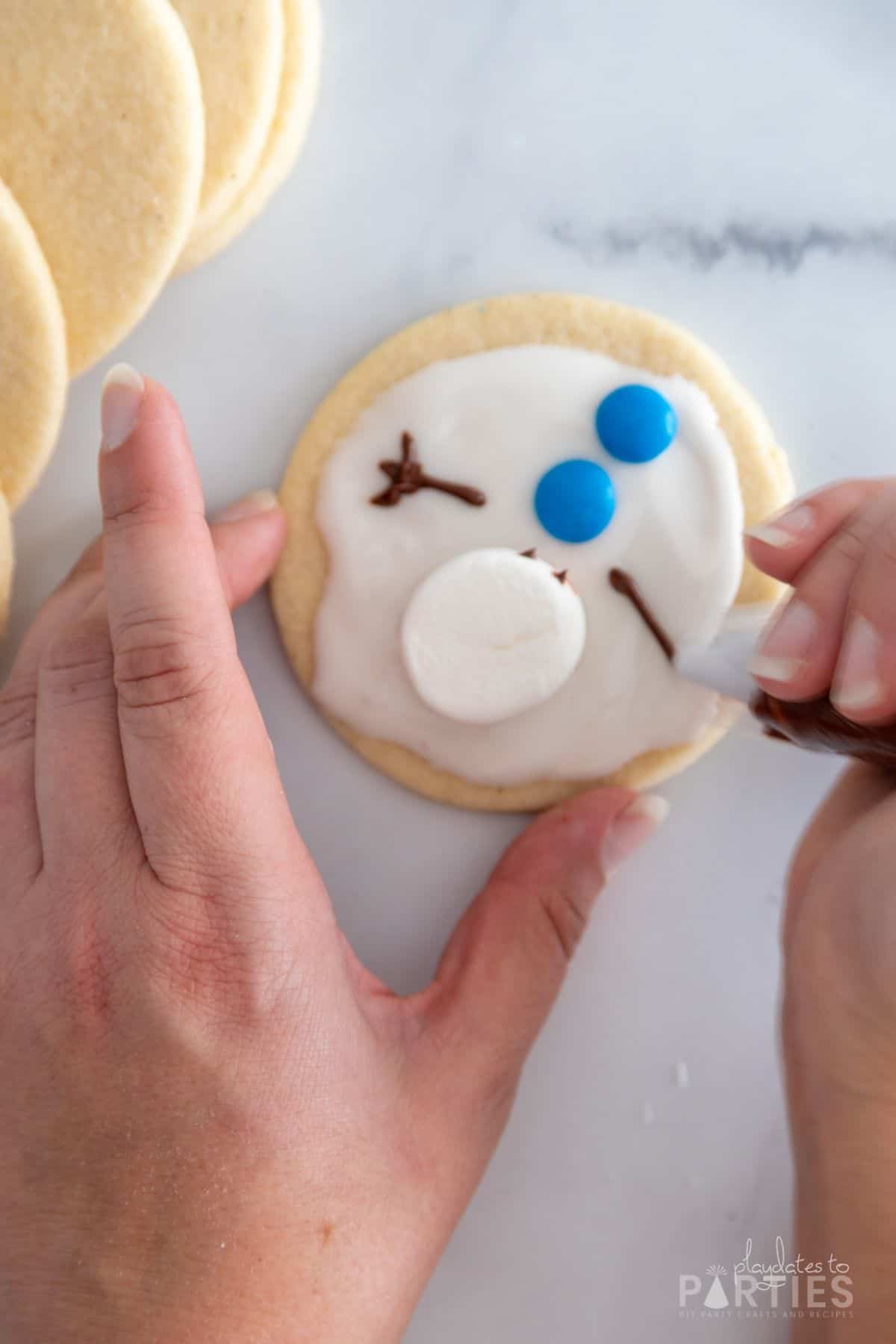 A woman's hands piping a chocolate snowman arm onto a cookie.