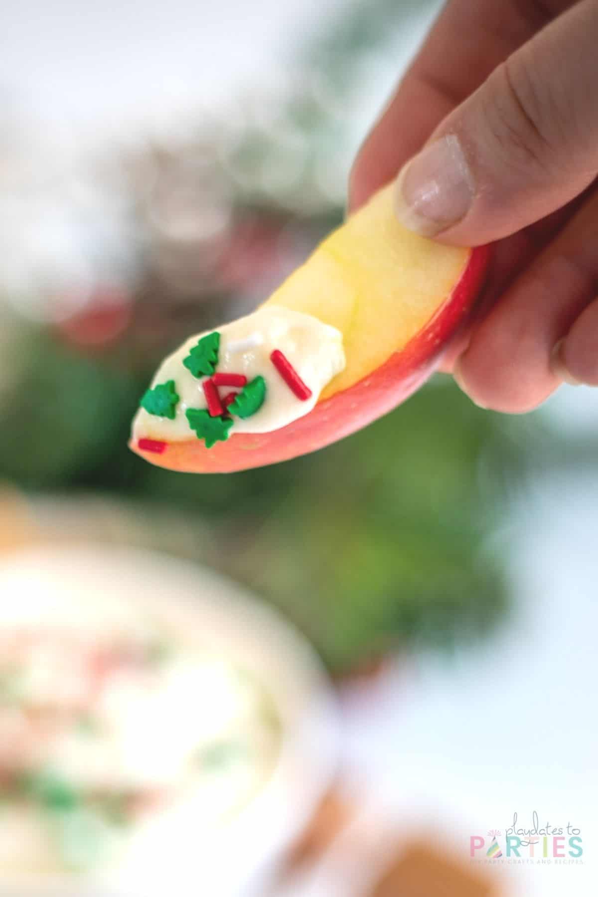 A woman's hand holding an apple slice with dip and sprinkles.