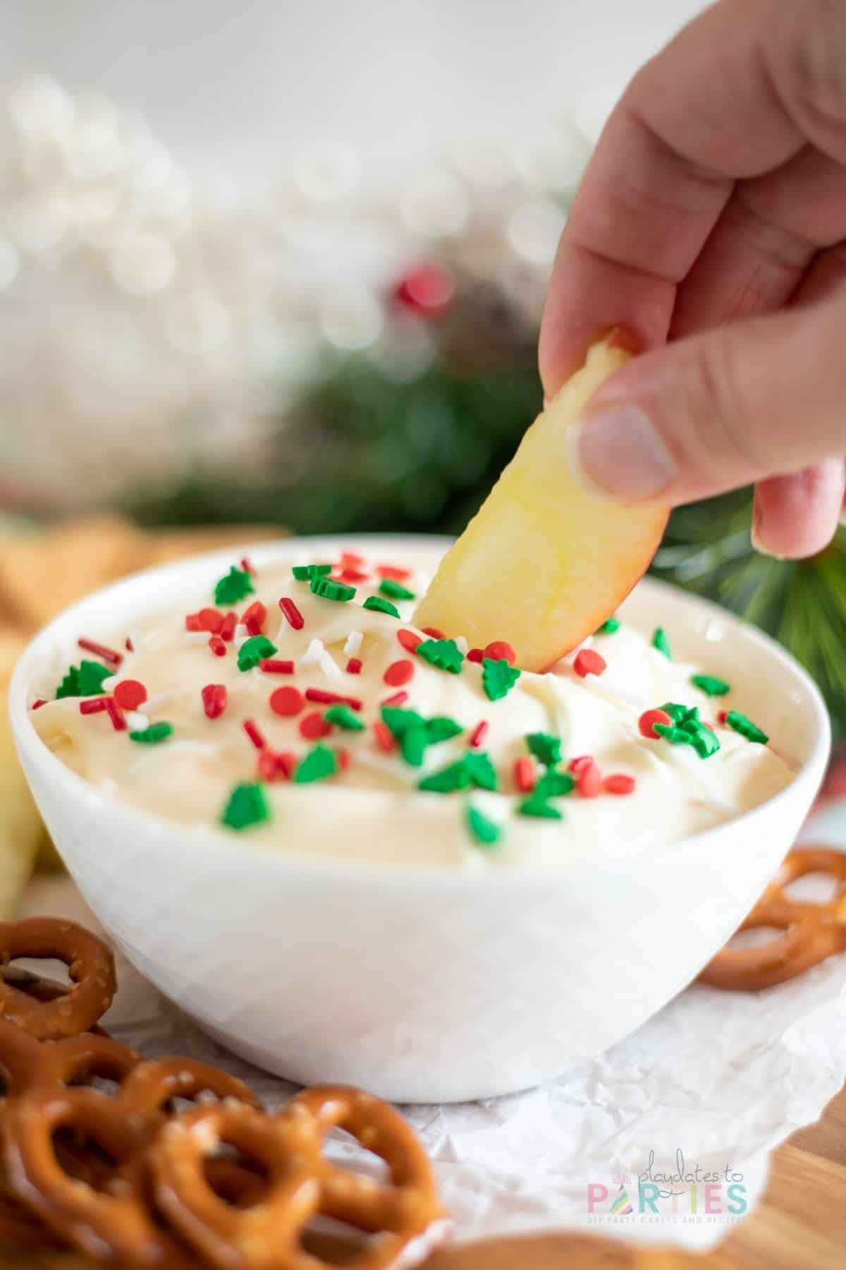 A woman's hand dipping an apple slice into a bowl of Christmas cookie dip.