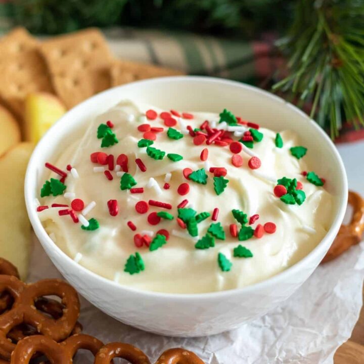 A bowl of sugar cookie dip surrounded by pretzels, fruit, and crackers.