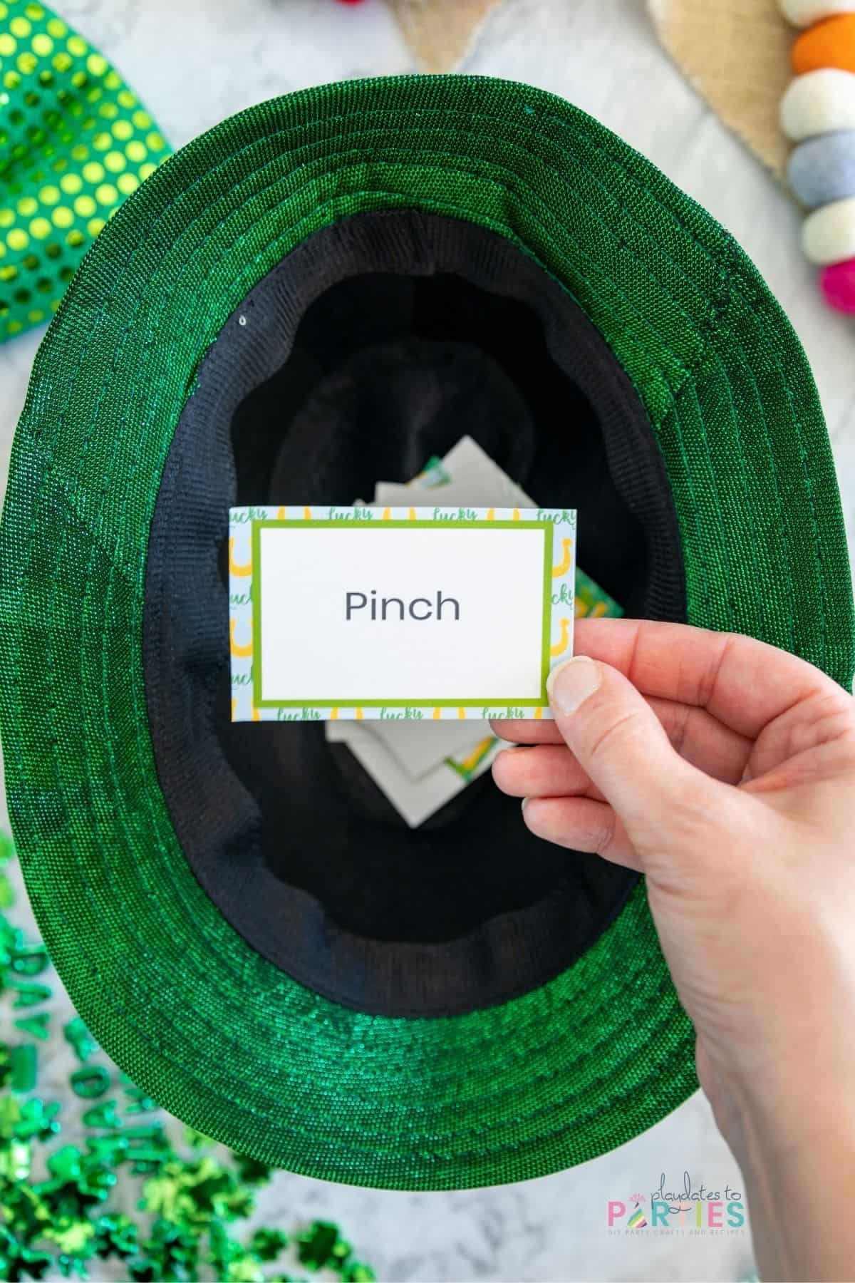 A woman's hand holding a St. Patrick's Day charades card over a green hat.