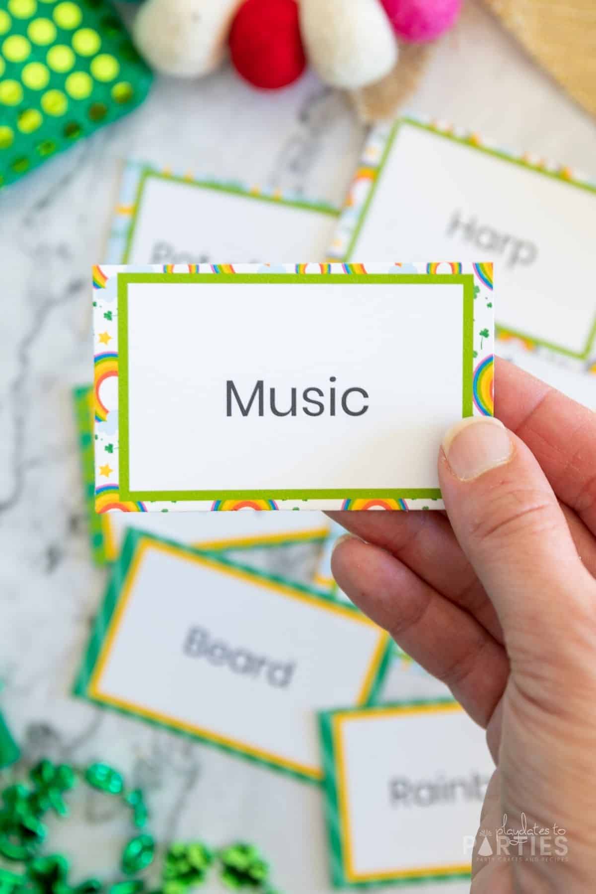 A hand holding a charades card with a rainbow border.