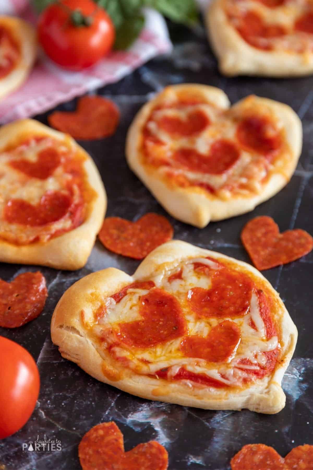 Mini heart shaped pizza on a dark surface with tomatoes scattered nearby.