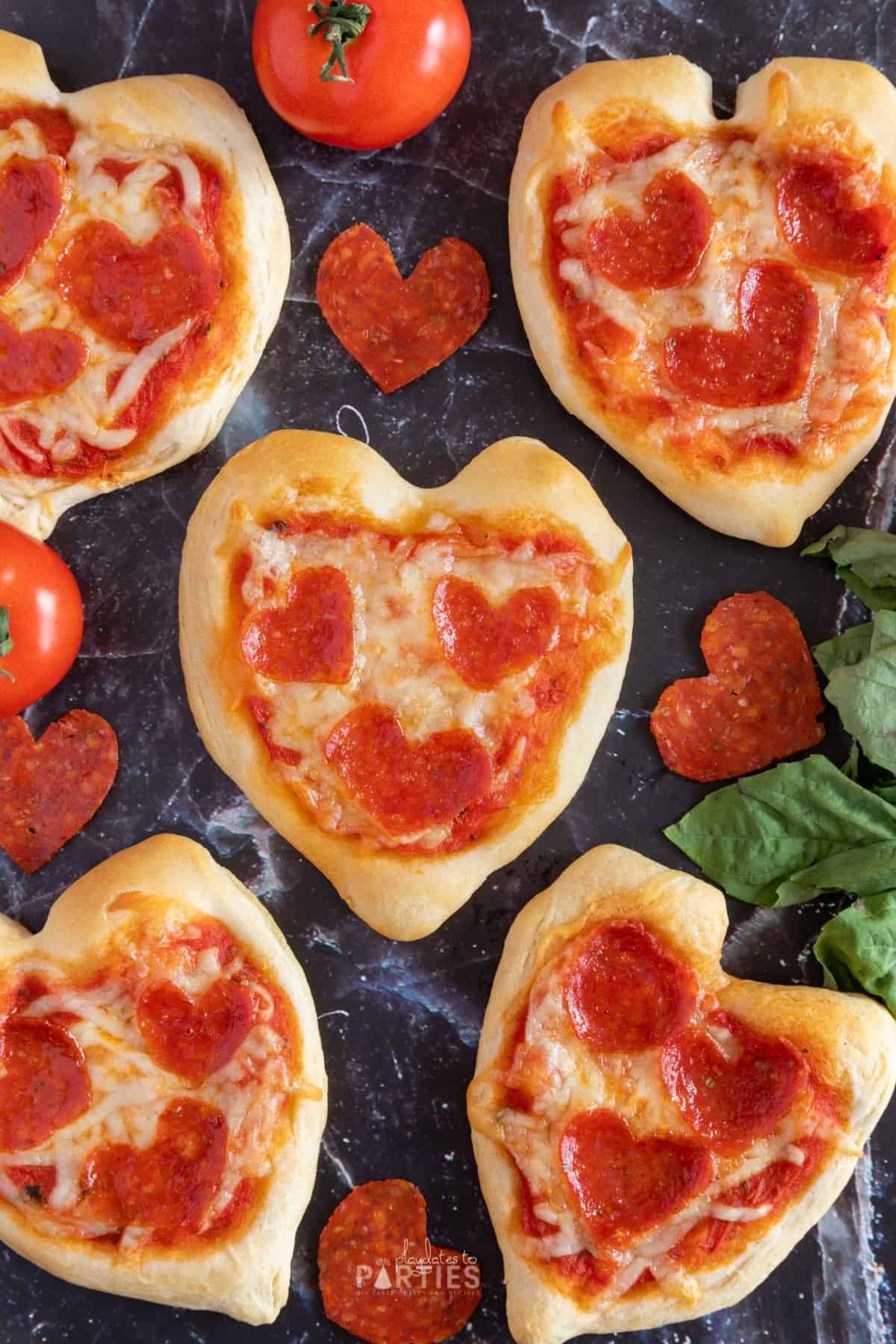 Overhead view of 5 mini heart shaped pizzas with heart shaped pepperoni and basil nearby.