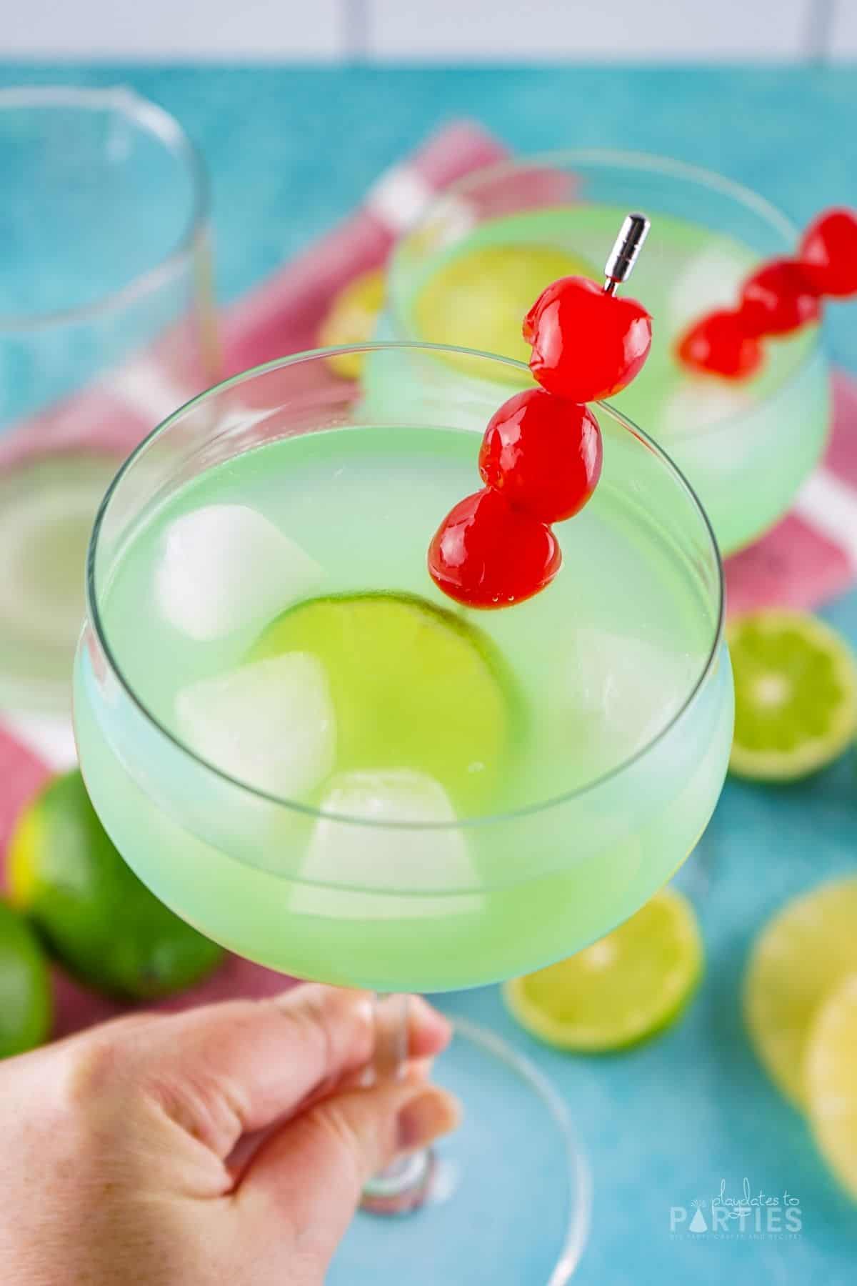 A woman's hand holding a green cocktail with lime and cherry garnish.