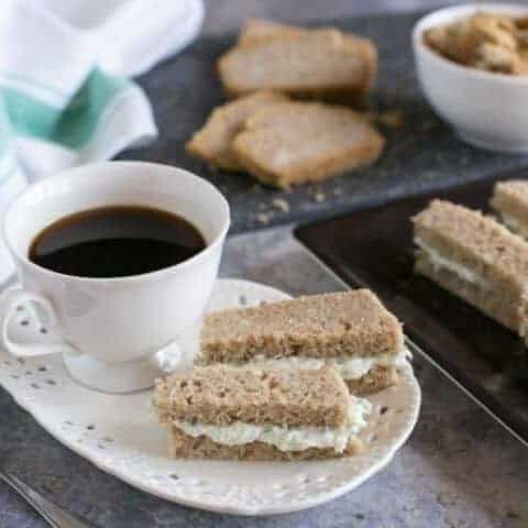 Two finger sandwiches on a plate next to a cup of coffee.