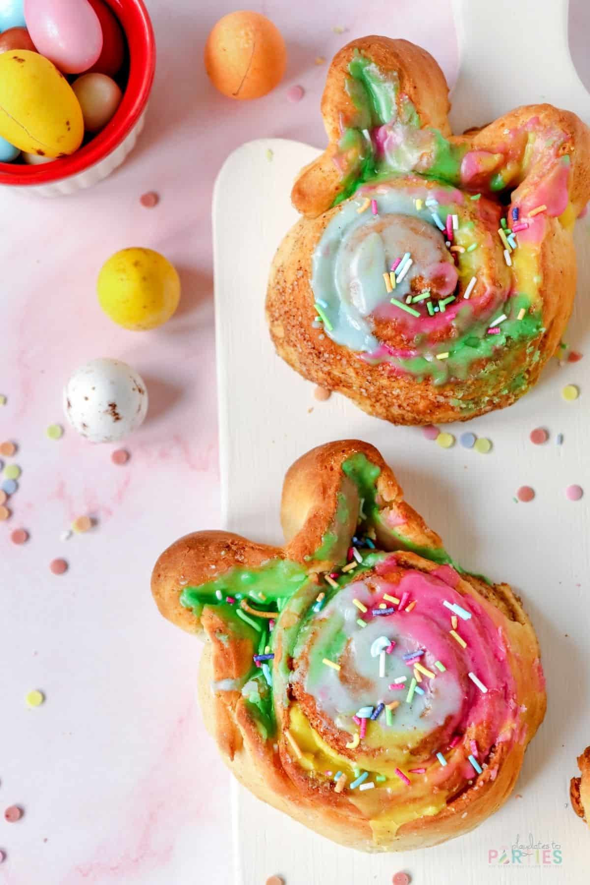 Bunny shaped cinnamon rolls with colorful icing and sprinkles on a cutting board.
