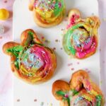 Four Easter bunny cinnamon rolls with colorful icing and sprinkles on a white cutting board.