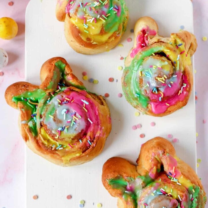 Four Easter bunny cinnamon rolls with colorful icing and sprinkles on a white cutting board.