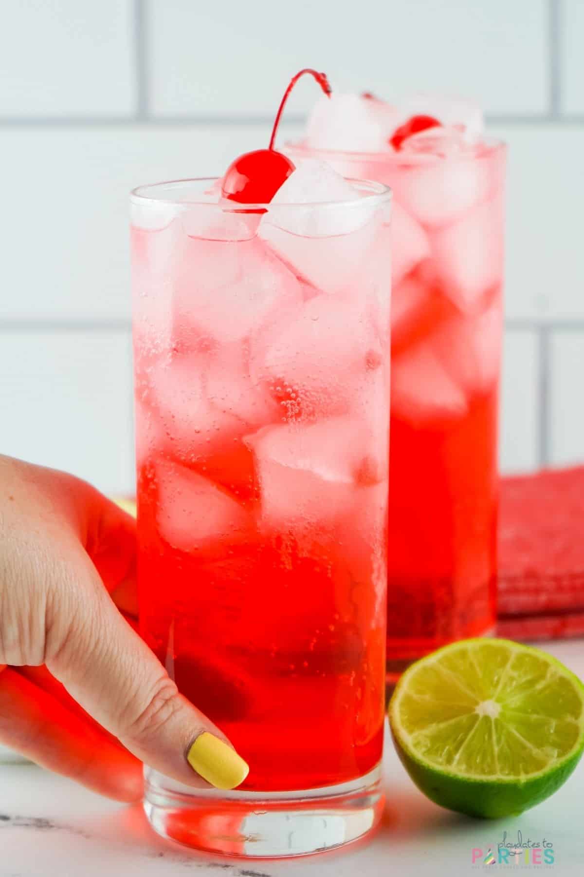 A woman's hand picking up a red drink garnished with a cherry.