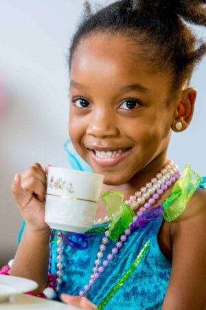 A little girl smiling and sipping from a teacup while playing a dress up tea party game.