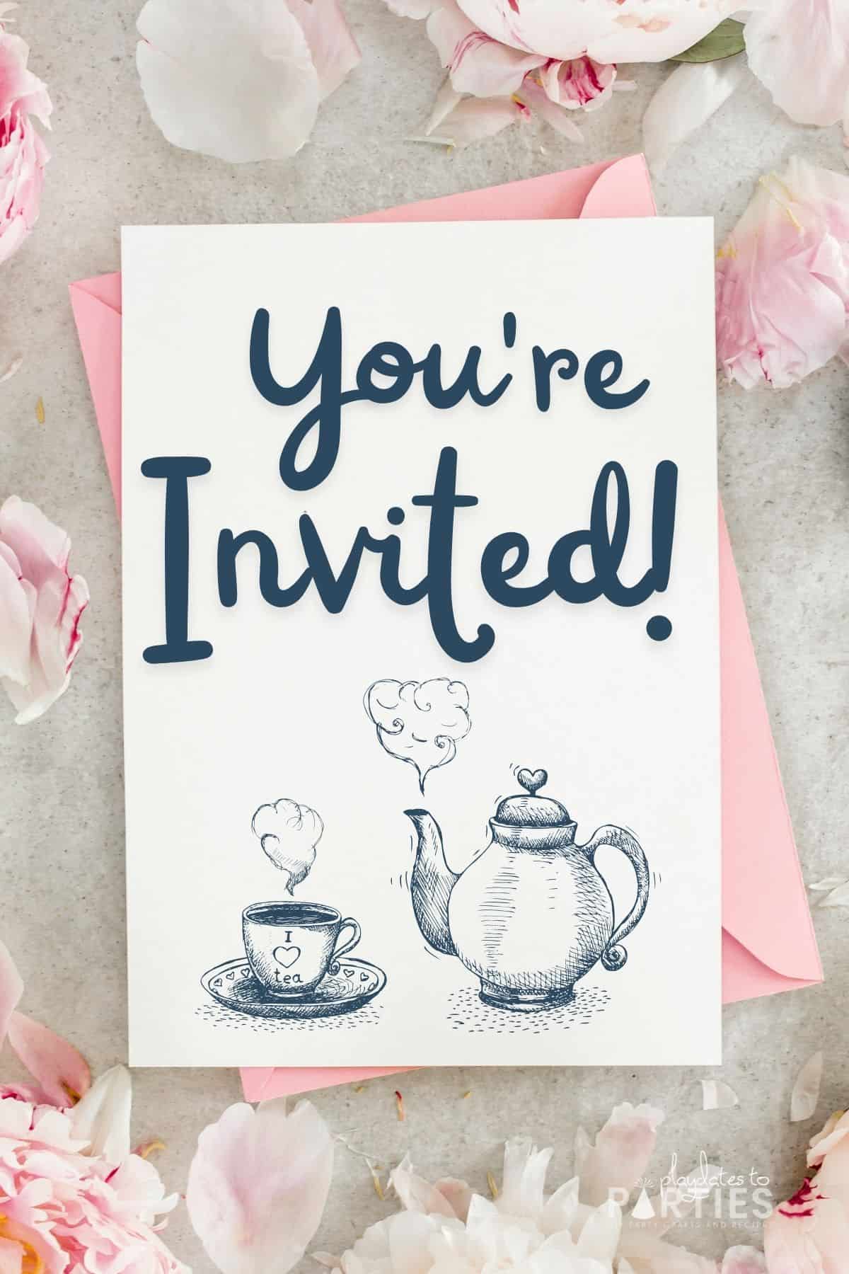 A tea party invitation with a tea pot surrounded by pink flowers on a gray surface.