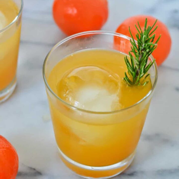 An orange whiskey cocktail garnished with fresh rosemary and surrounded by tangerines.