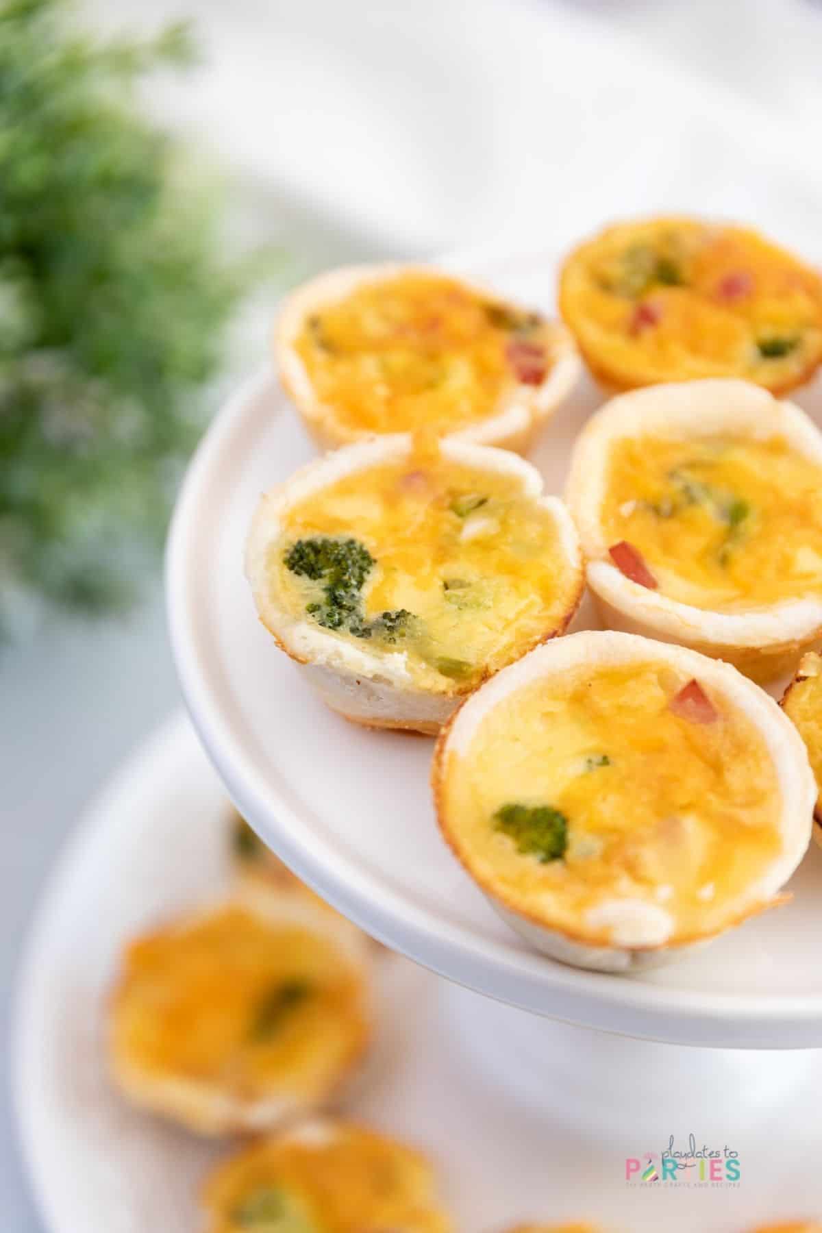 Mini ham and broccoli quiche appetizers on a white cake stand for a party.