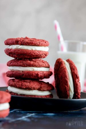 3 red velvet sandwich cookies stacked on a plate in front of a glass of milk.