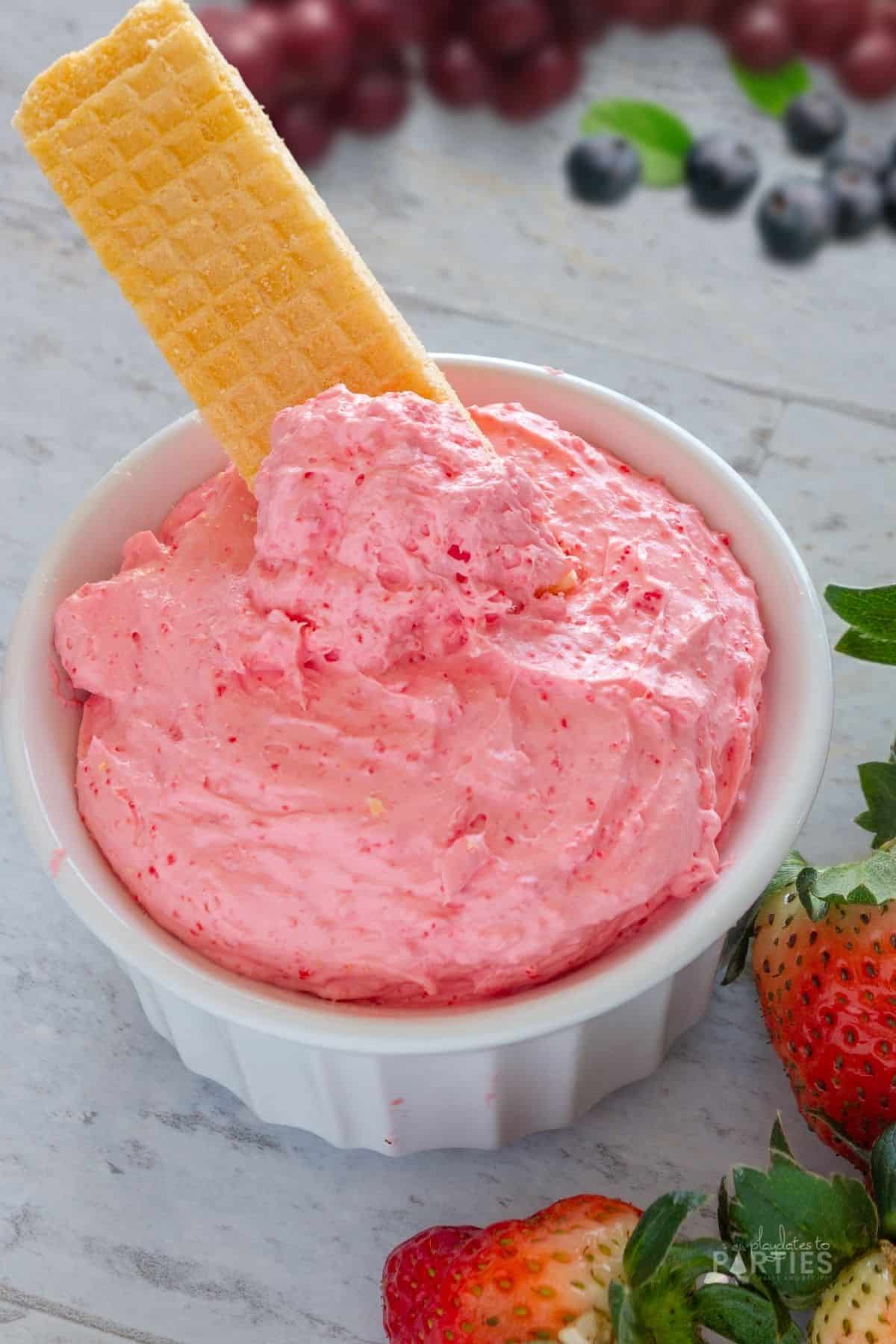 A sugar wafer sitting in a bowl of pink strawberry fruit dip with cream cheese.