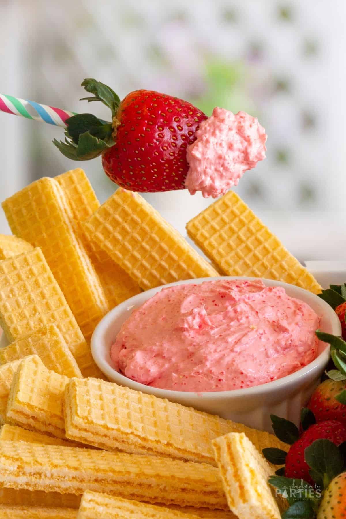 A strawberry with pink fruit dip over a bowl filled with sugar wafers and fruit.