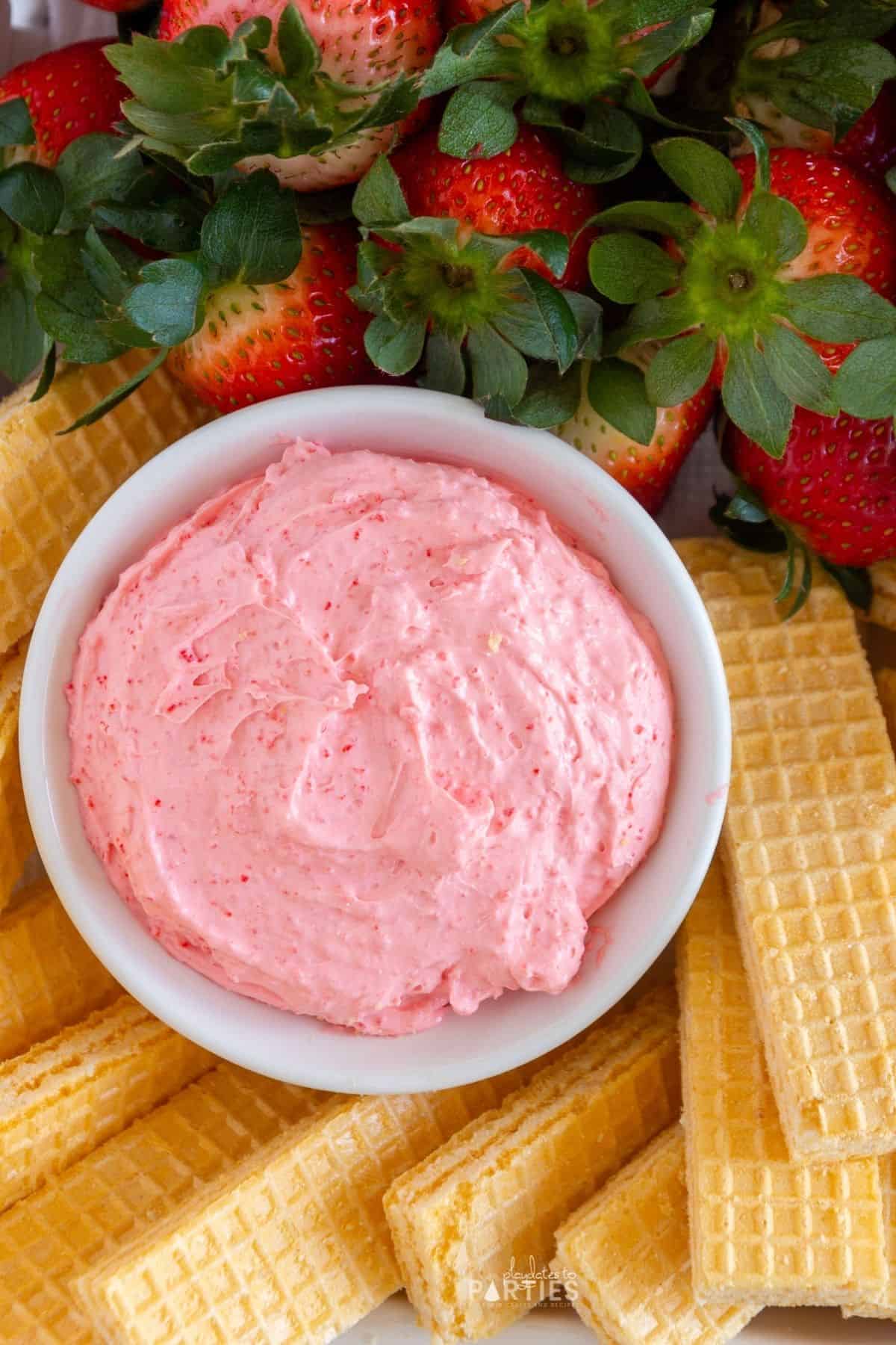 A bowl of pink dip surrounded by sugar wafers and fresh fruit.