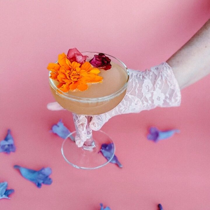 A feminine gloved hand holding a cocktail that is garnished with fresh flowers.
