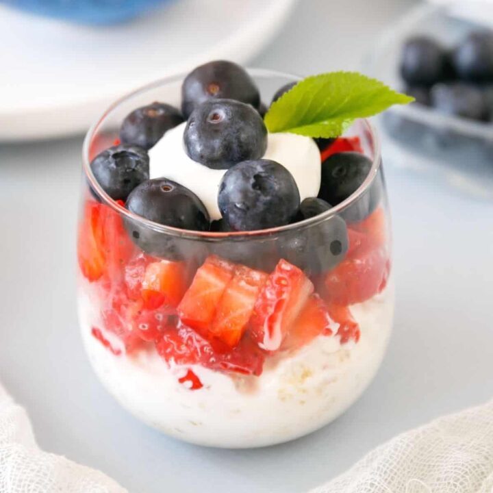 Parfait with greek yogurt, granola, strawberries, and blueberries on a blue surface.