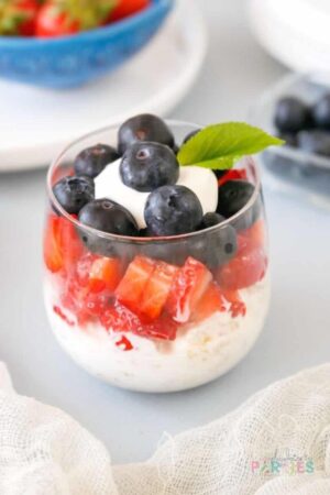 Parfait with greek yogurt, granola, strawberries, and blueberries on a blue surface.