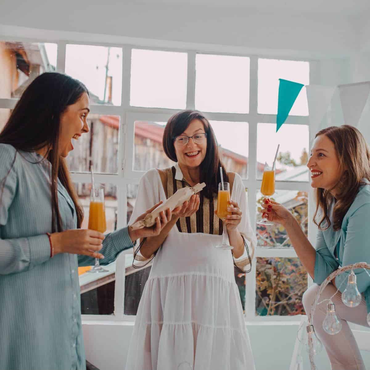 Three women smile and giggle at a baby shower while passing around a wrapped gift.