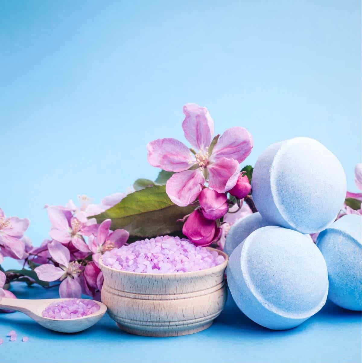 A tub of bath salts with fresh flowers and bath bombs nearby.