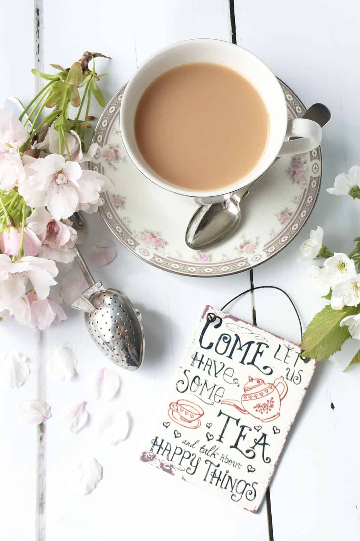 A cup of tea on a white wood surface with a sign that says come let us have som tea.