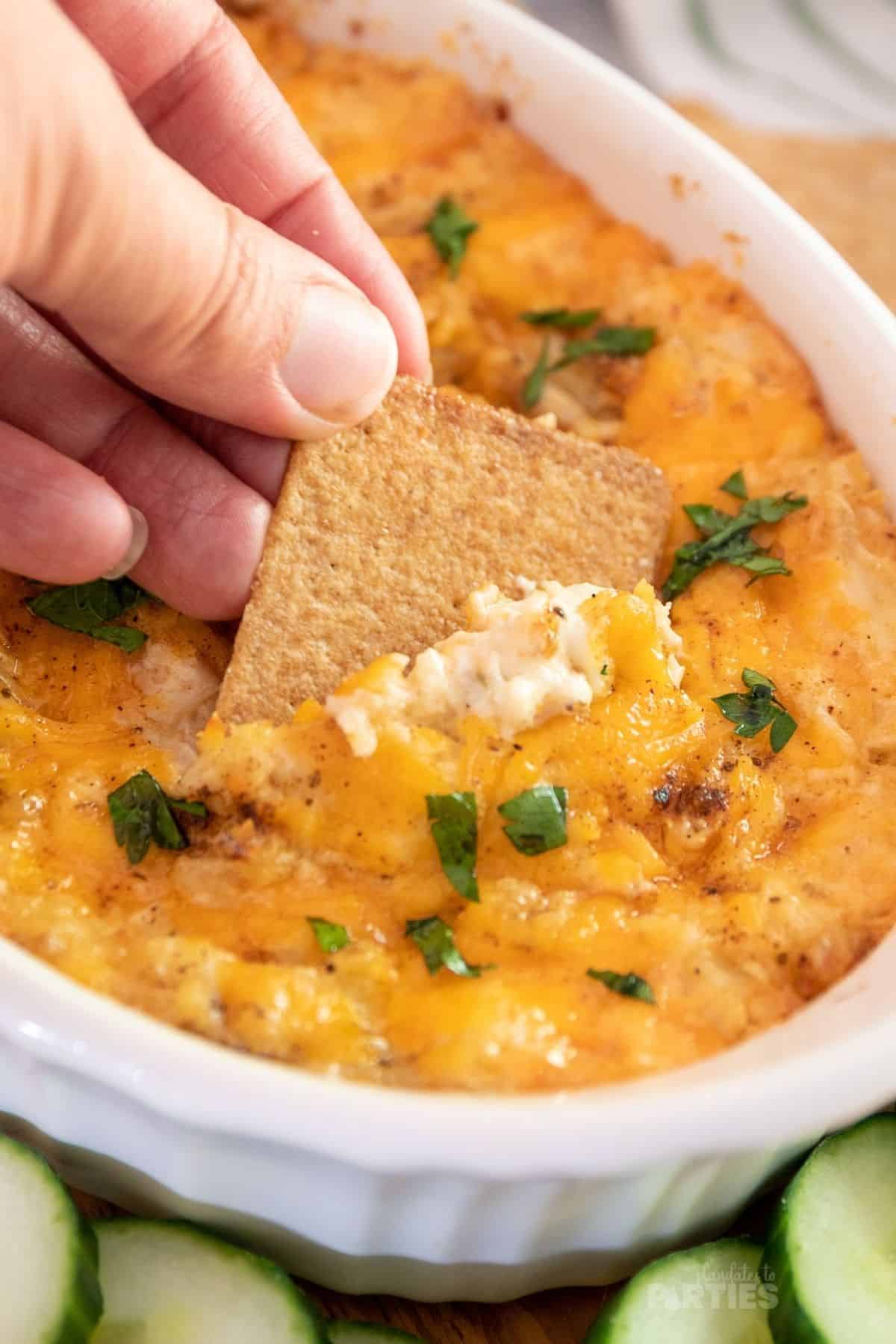 A woman's hand dipping a cracker into a bowl of cheesy crab dip.