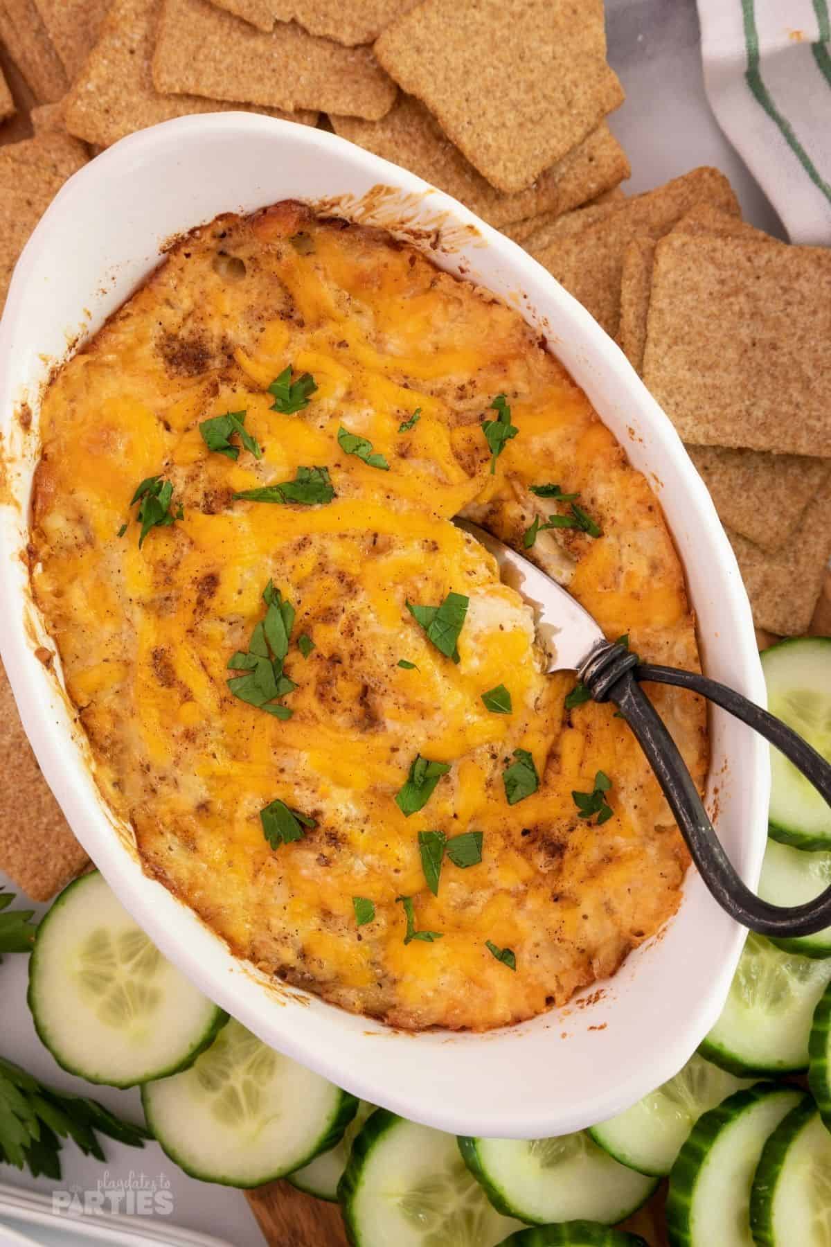 Top down view of a bowl of warm crab dip with crackers and cucumber slices.