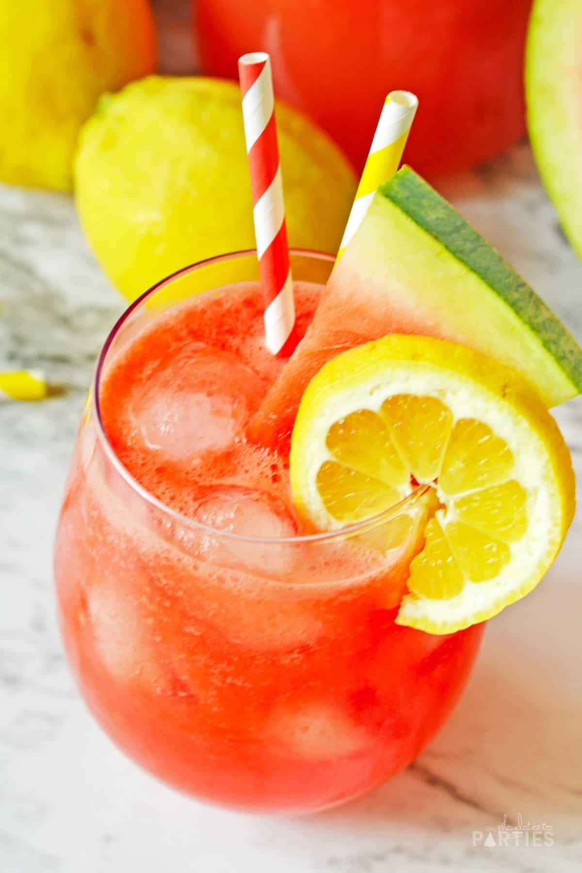 Overhead view of a red juice drink garnished with a lemon slice, watermelon wedge and colorful paper straws.