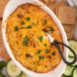 Top down view of a bowl of warm crab dip with crackers and cucumber slices.