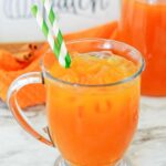 A mug of pumpkin juice recipe with green paper straws with cinnamon sticks, a pumpkin patch sign, and a pitcher of juice in the background.