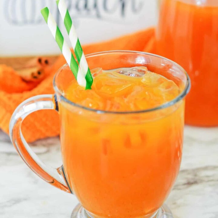 A mug of pumpkin juice recipe with green paper straws with cinnamon sticks, a pumpkin patch sign, and a pitcher of juice in the background.