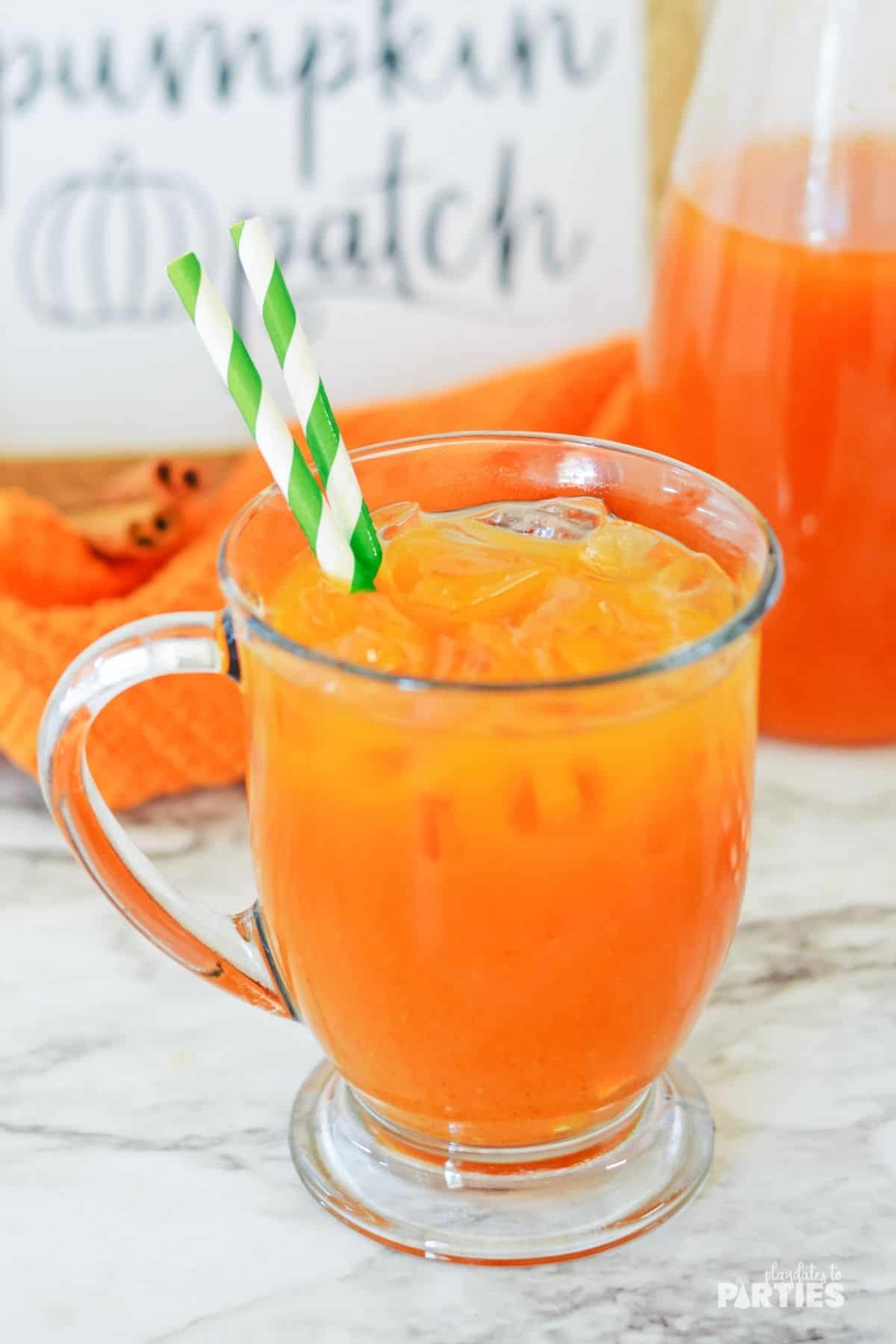 A mug of pumpkin juice recipe with green paper straws with cinnamon sticks, a pumpkin patch sign, and a pitcher of juice in the background.