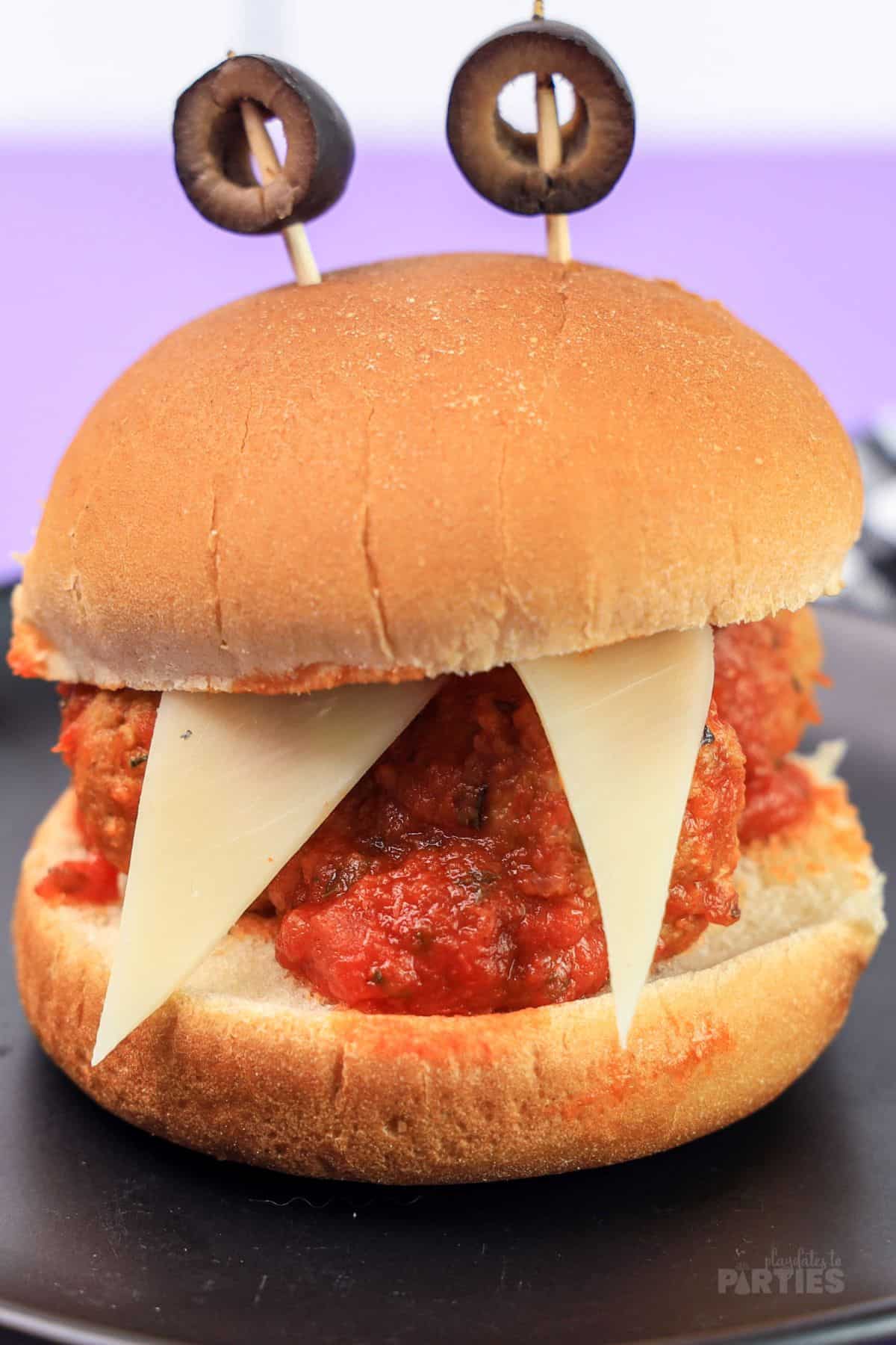 Two triangle cheese teeth stick out of a monster meatball slider with olives on top for eyes.