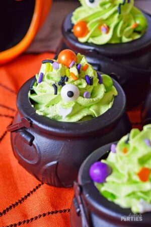 Halloween pudding cups in mini witch cauldrons on an orange tablecloth.