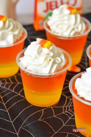 Plastic cups with layers of yellow and orange jello with whipped cream and candy corn.