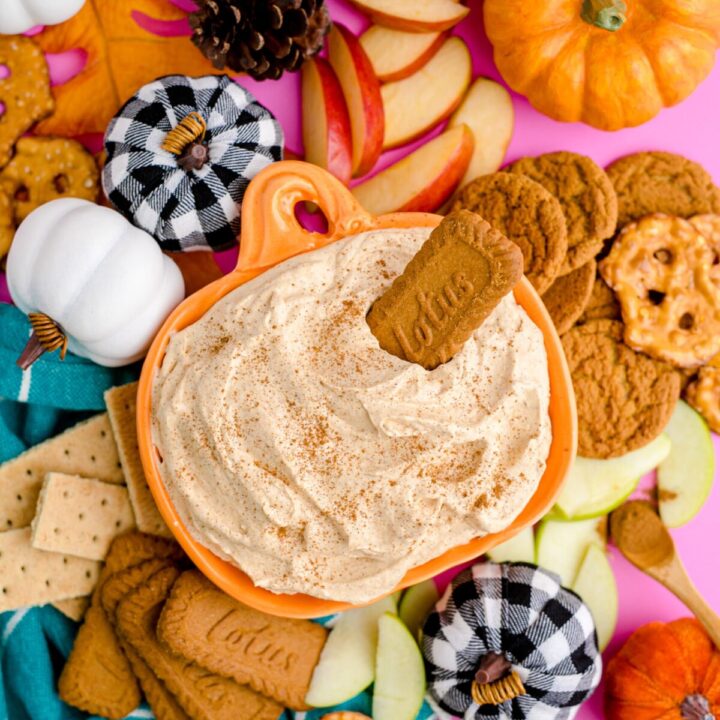 A pumpkin shaped bowl is filled with pumpkin spice dip and is surrounded by cookies, and fresh fruit for dipping.