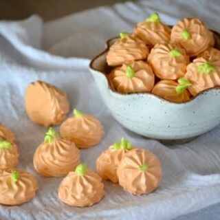 Three dimensional pumpkin spice meringue cookies scattered on a towel and placed in a bowl.