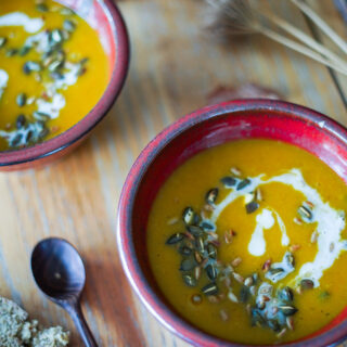 A red bowl filled with pumpkin soup that is garnished with coconut cream and pumpkin seeds.