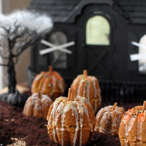 Mini bundt cakes are stacked and decorated to look like little pumpkins in front of a haunted house.