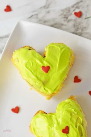 Two heart shaped grinch cinnamon rolls with green frosting and heart sprinkles.